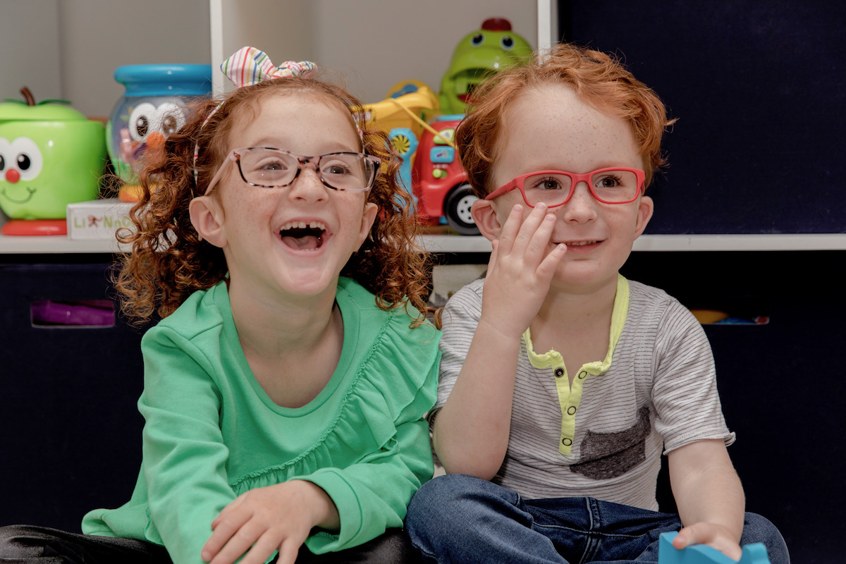 Children wearing glasses, one in pink frames and the other in red frames, smiling and laughing.