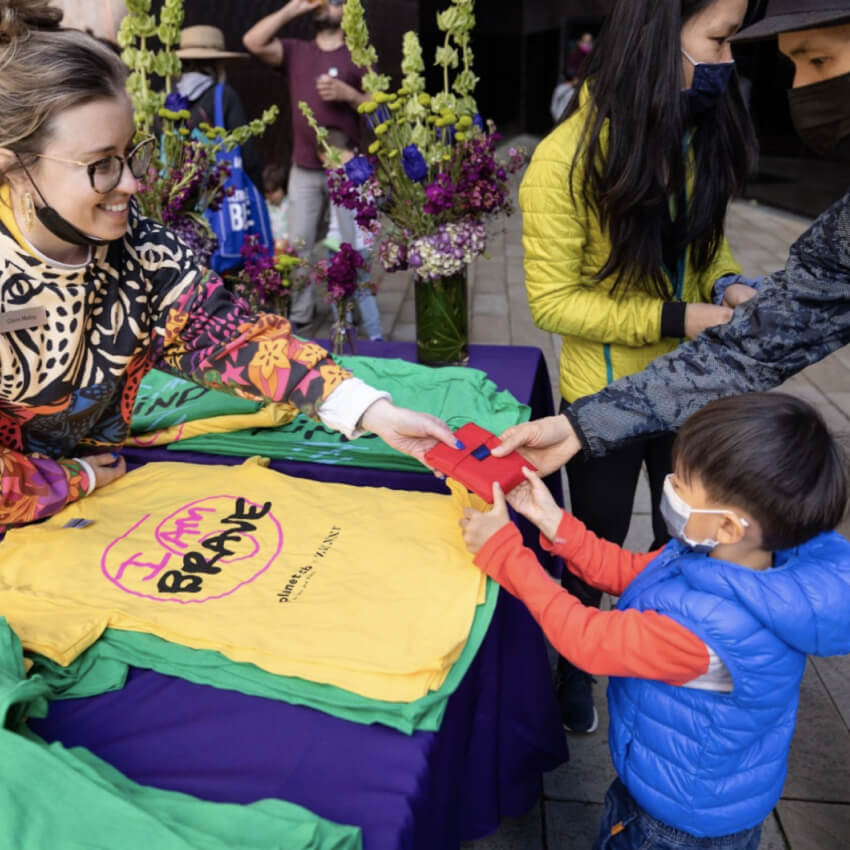 T-shirts with "I AM BRAVE" printed on them and a child holding a red gift bag being handed over.