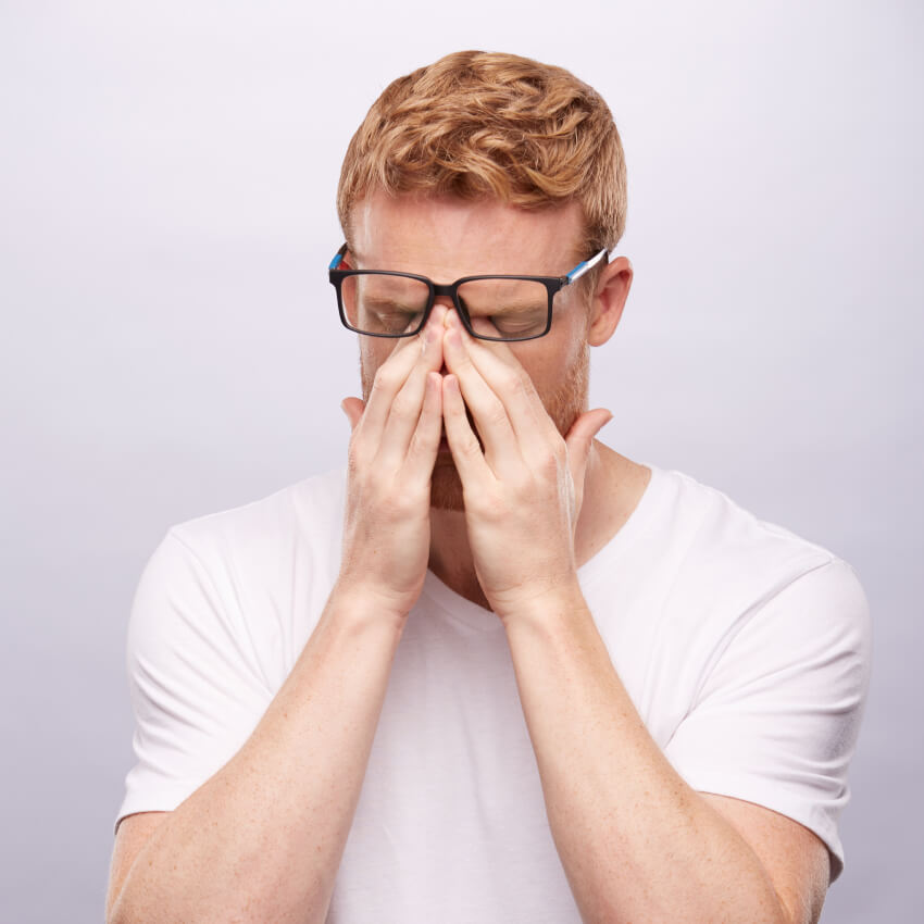 Man wearing glasses and white T-shirt pinching the bridge of his nose, appearing stressed or tired.