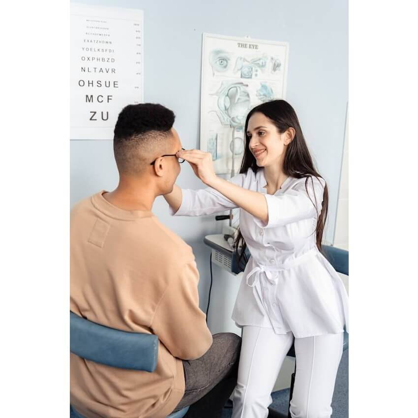 Optometrist adjusts eyeglasses on patient. Eye chart in the background reads: "EKATZHQWV YOELKSFDI OXPHBZD NLTAVR OHSUE MCF ZU."