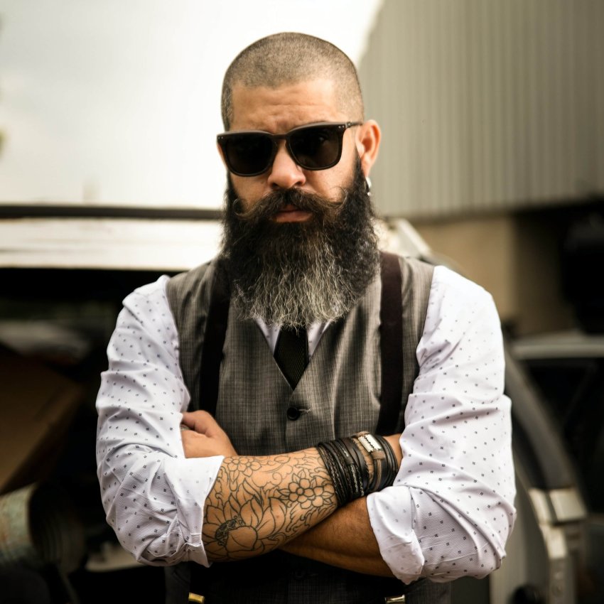 Bearded man in sunglasses wearing a vest with tie, rolled-up sleeves, and wristbands, arms crossed.
