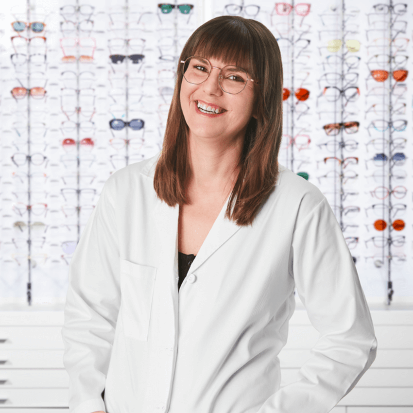 Smiling optician wearing glasses and a white coat, standing in front of a wall display of various eyeglasses.