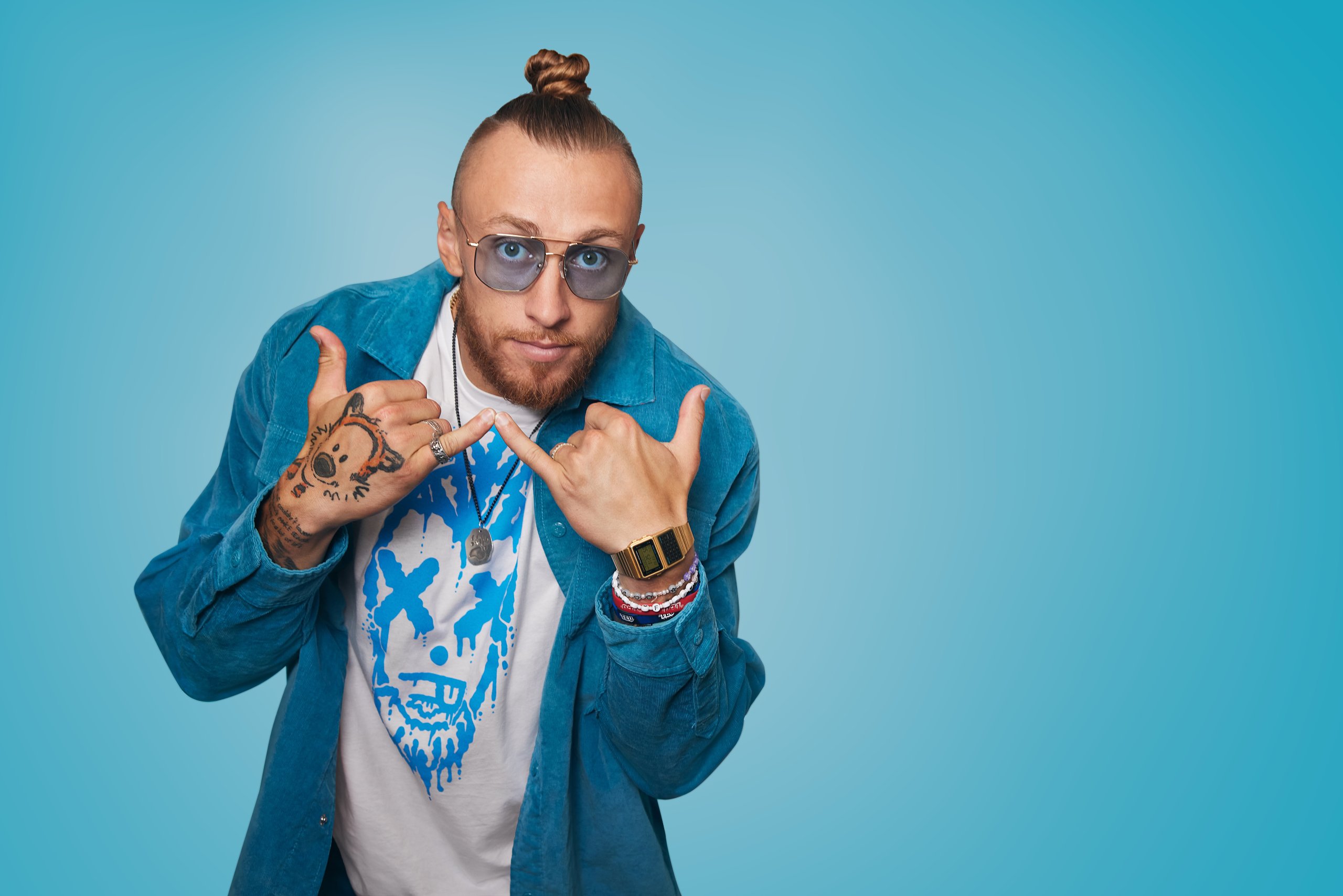Man wearing blue jacket and white t-shirt with blue illustrated face. He sports sunglasses and a top knot.