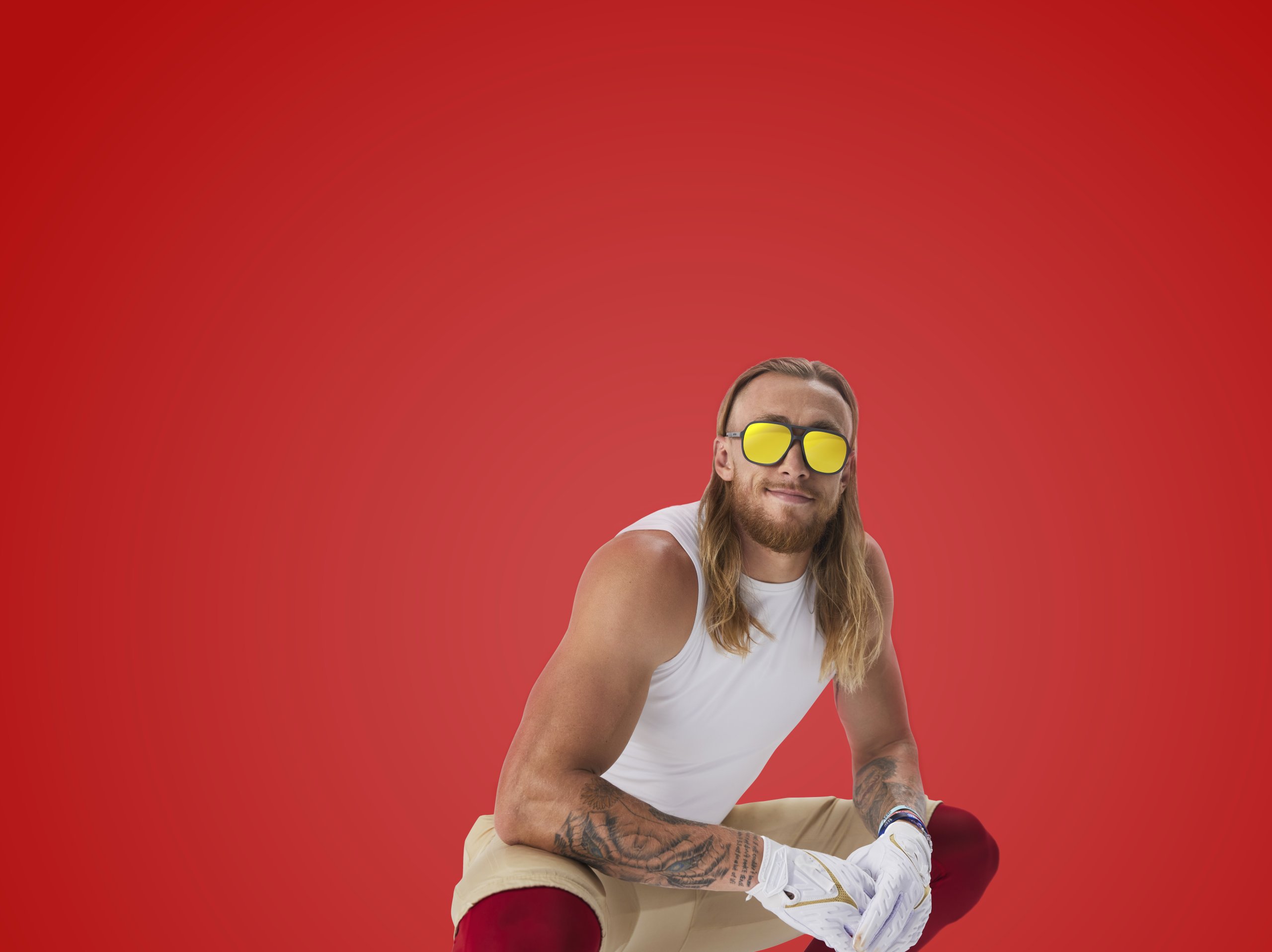 Person wearing yellow reflective sunglasses, a white sleeveless shirt, and white gloves against a red background.