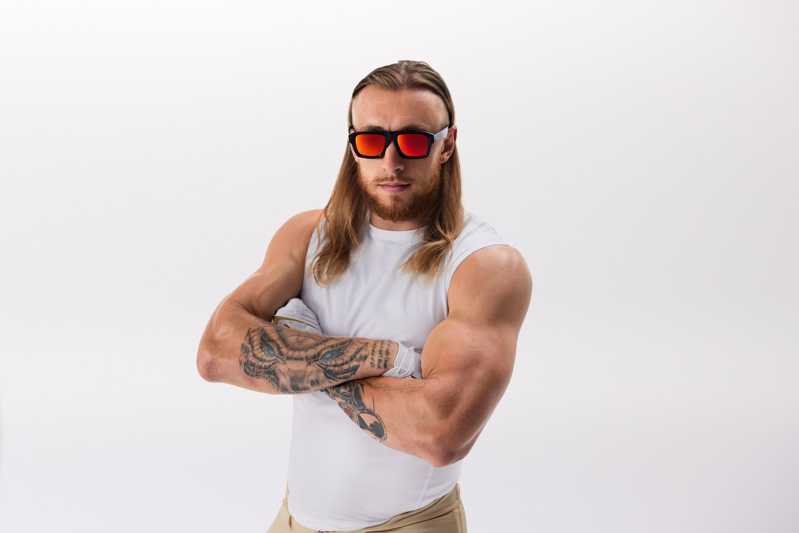 Man wearing red-lens sunglasses, a white sleeveless shirt, showcasing arm tattoos with a confident pose.