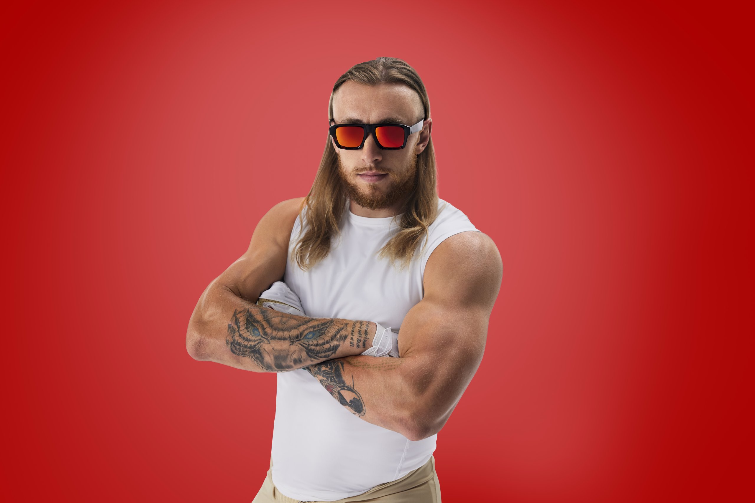Man wearing black and red sunglasses with reflective lenses, a white sleeveless shirt, and displaying arm tattoos.