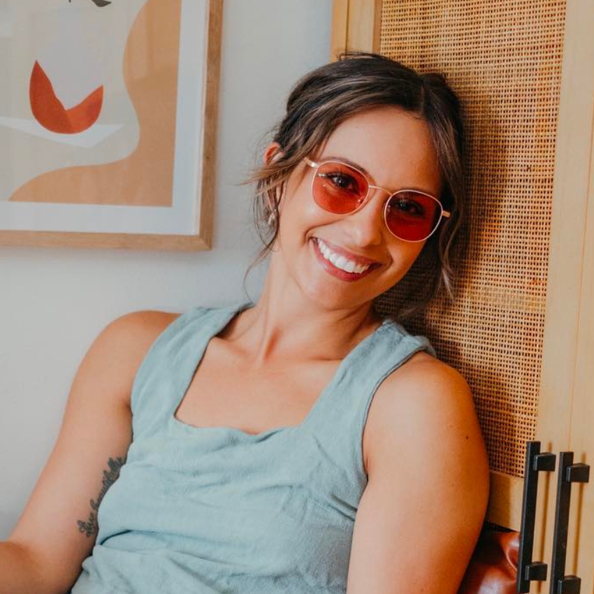 Woman smiling and wearing red-tinted sunglasses with a light blue sleeveless top.