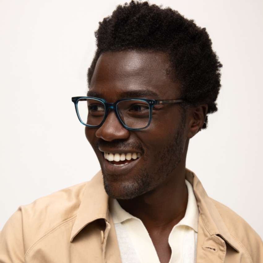 Man wearing black-framed eyeglasses, beige coat, white shirt, smiling and looking to the side.