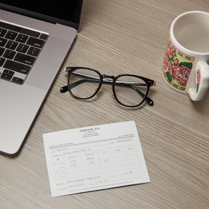 Black-rimmed glasses on a wooden desk next to a prescription card and a laptop. Card text: JOHN DOE, O.D. 123 Anywhere St. New York, NY XXXXX Telephone: (555) XXX-XXXX Lic #: XXXXTXXX Date: 04/08/2018 For