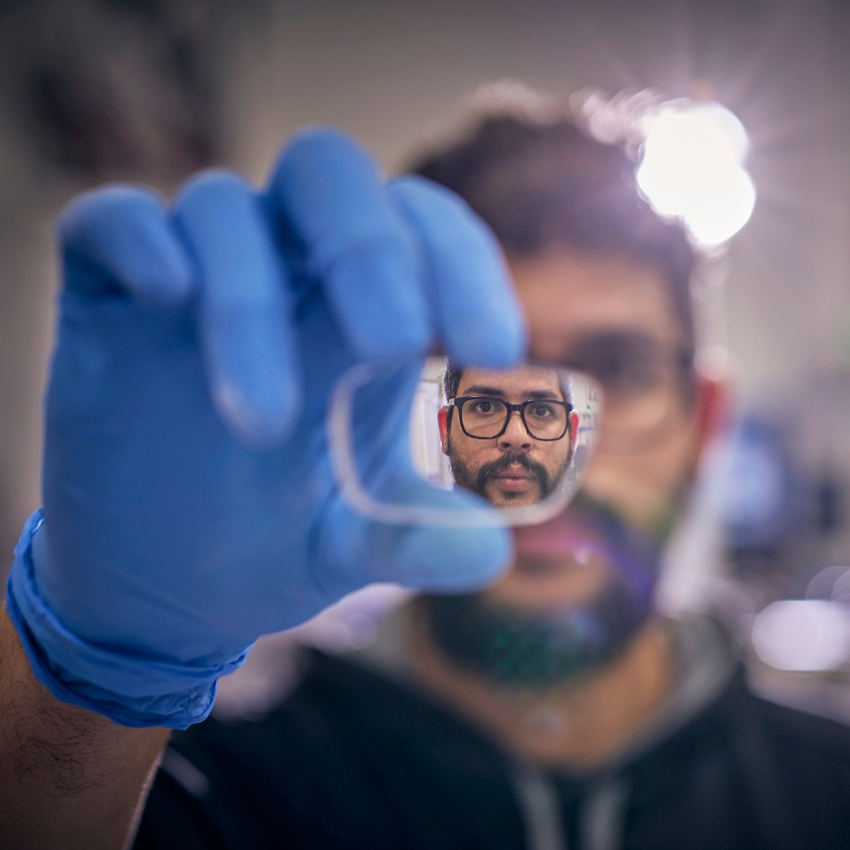 Close-up of a person wearing blue gloves holding a lens, reflecting their face with glasses.