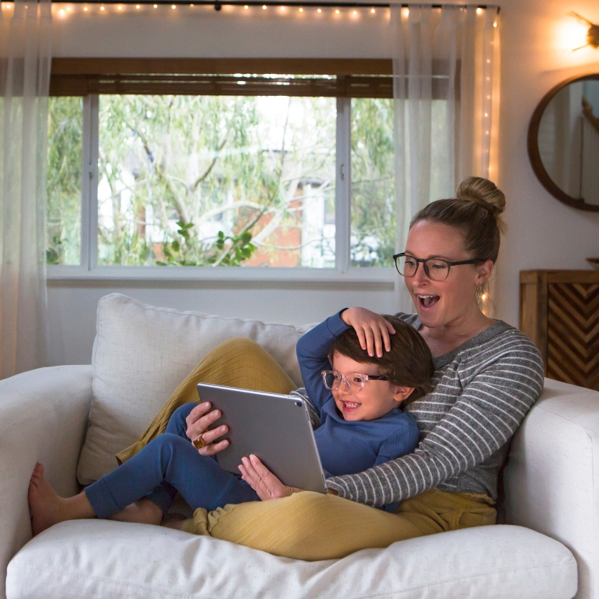 Adult and child wearing glasses, sitting on a couch, smiling and looking at a tablet screen together.