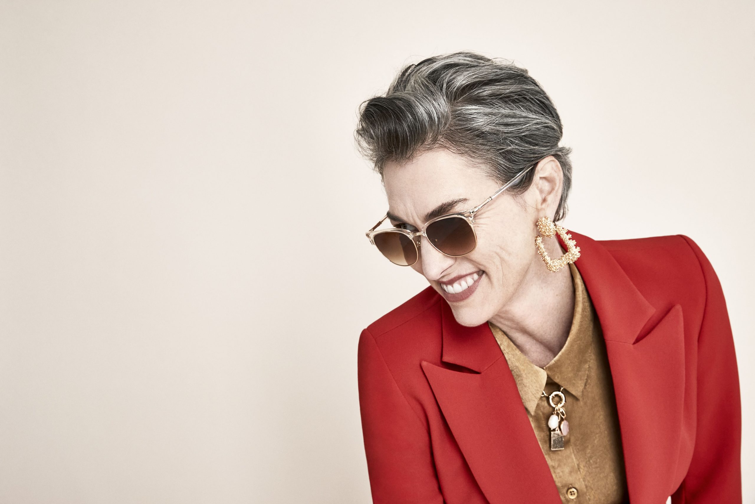 Woman wearing gold drop earrings, sunglasses, a red blazer, and a tan shirt with a necklace.