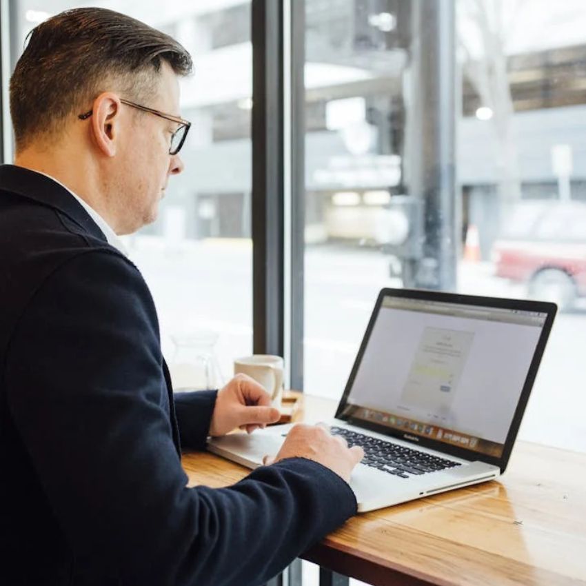Man working on a MacBook laptop at a wooden desk by a window