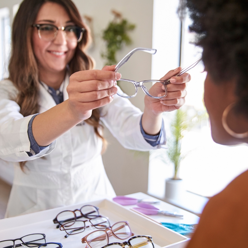 Optician holding a pair of eyeglasses towards a customer, with multiple eyeglass frames on the table.