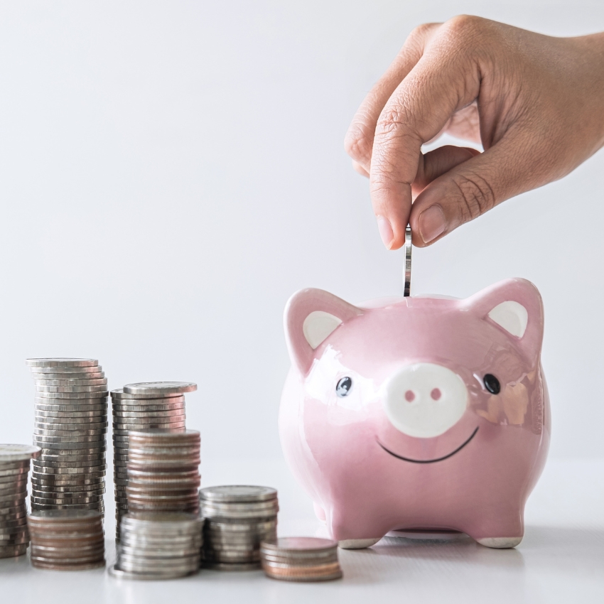 Hand placing a coin into a smiling pink piggy bank next to stacks of coins.