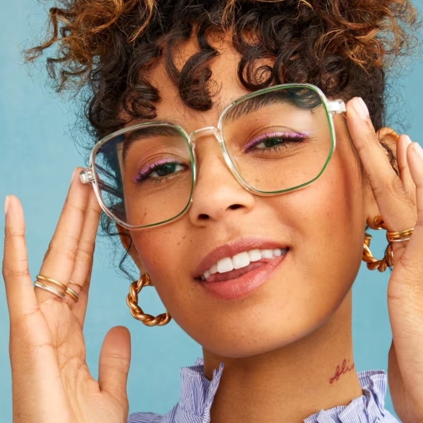 Woman wearing oversized green-rimmed glasses with her hands touching the frames. She also has gold hoop earrings and rings.