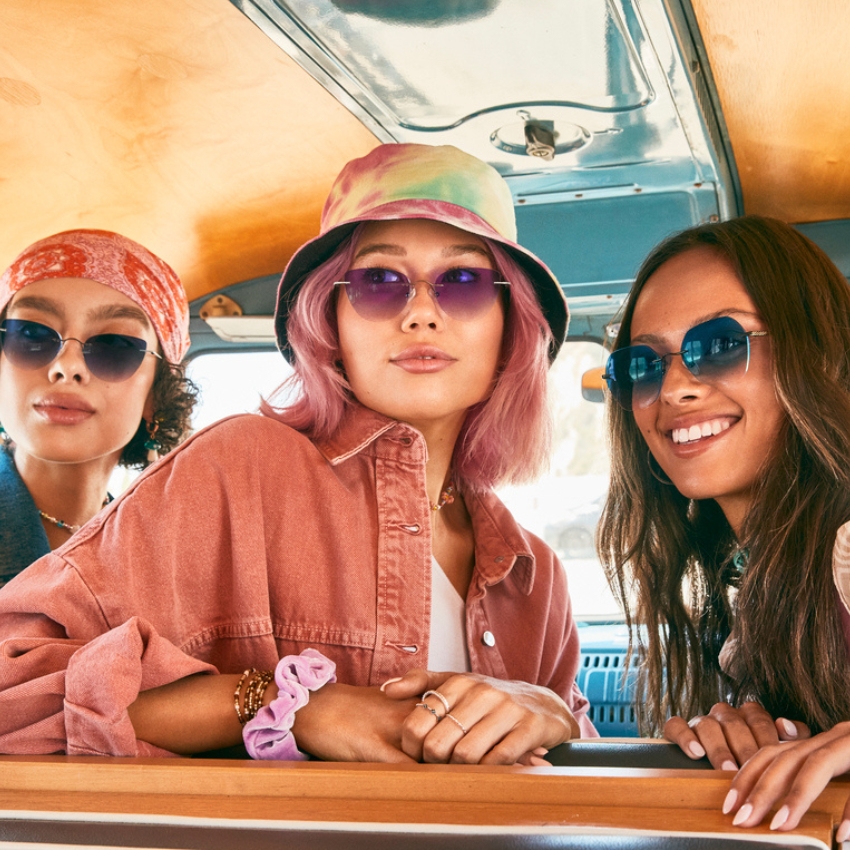 Three women wearing sunglasses and casual clothing, with one in a pink jacket and bucket hat at the center.