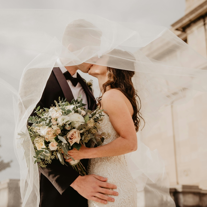 Bride and groom kissing under veil. Bride holds bouquet of pale roses and white flowers with eucalyptus.