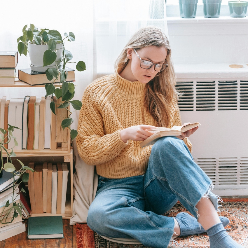 Person wearing a mustard yellow knit sweater and blue jeans, reading a book.