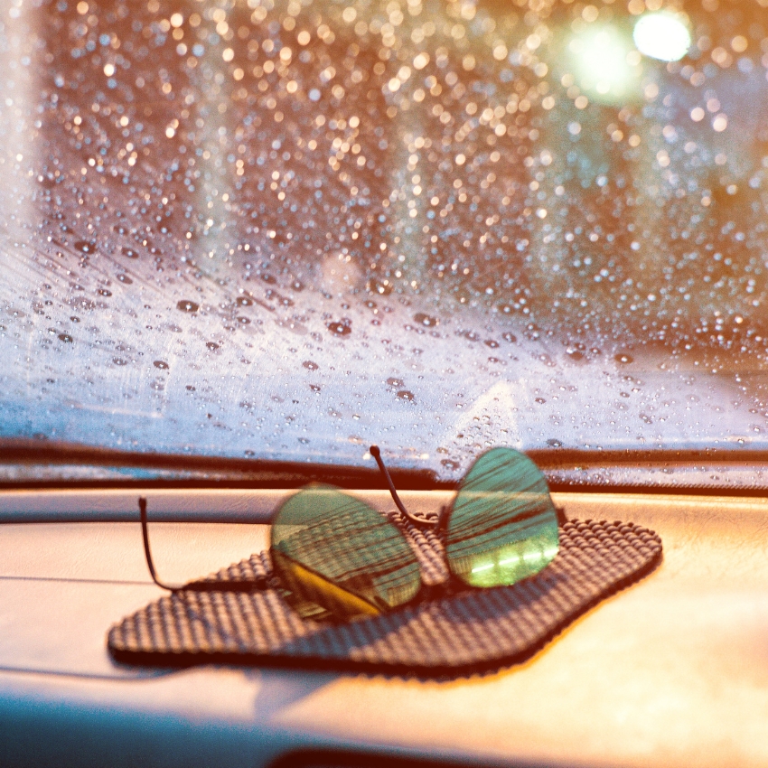 Green-tinted aviator sunglasses on a textured mat on a car dashboard, with a raindrop-covered windshield.
